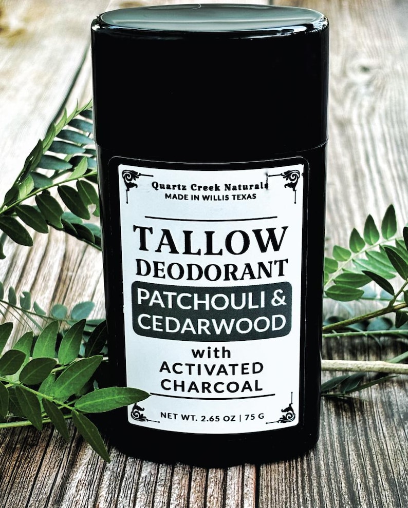 K&ouml;nnte beinhalten: Ein schwarzer zylindrischer Deodorant-Beh&auml;lter mit einem wei&szlig;en Etikett mit der Aufschrift Quartz Creek Naturals, Tallow Deodorant, Patchouli & Cedarwood with Activated Charcoal und Net Wt. 2.65 oz | 75 g. Das Produkt steht aufrecht neben gr&uuml;nen Bl&auml;ttern.