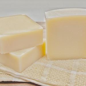 May include: Three rectangular bars of solid, off-white soap with smooth, matte surfaces. Two bars are stacked horizontally on the left, while one bar stands vertically on the right. The soap bars are resting on a folded, light beige checkered cloth.