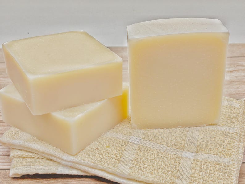 May include: Three rectangular bars of solid, off-white soap with smooth, matte surfaces. Two bars are stacked horizontally on the left, while one bar stands vertically on the right. The soap bars are resting on a folded, light beige checkered cloth.