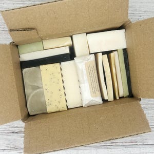 May include: An open cardboard box filled with various handmade soaps. The soaps are different shapes, sizes, and colors, including white, black, green, and yellow. Some soaps have visible seeds or textures. A wrapped soap bar is also visible.