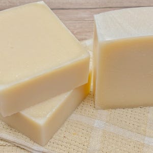 May include: Three rectangular bars of solid, off-white soap with a smooth, matte finish. The bars are stacked and positioned together, displaying a uniform, light cream color throughout each piece, showcasing a simple and clean appearance.