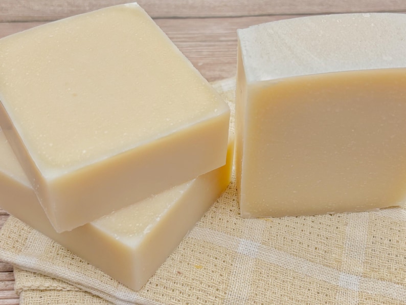 May include: Three rectangular bars of solid, off-white soap with a smooth, matte finish. The bars are stacked and positioned together, displaying a uniform, light cream color throughout each piece, showcasing a simple and clean appearance.