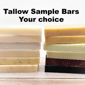 May include: A stack of eight different scented soap bars, including a black bar, a brown bar, a white bar, a light green bar, a yellow bar, and a few beige bars. The text "Tallow Sample Bars Your choice" is written above the bars.