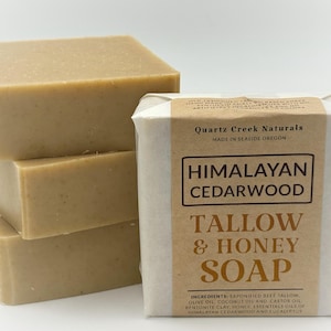 May include: Four rectangular bars of soap stacked next to a wrapped bar. The unwrapped bars are light brown. The wrapped bar has a brown label with the text "Himalayan Cedarwood Tallow & Honey Soap."
