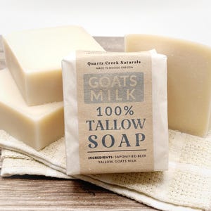 May include: Four rectangular bars of off-white soap. One bar is wrapped in a tan paper label with black text reading Quartz Creek Naturals, Goats Milk, 100% Tallow Soap, and Ingredients: Saponified Beef Tallow, Goats Milk. The soap is stacked.