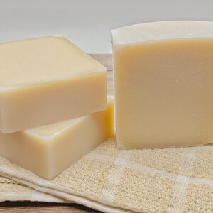 May include: Three rectangular bars of solid, off-white soap with smooth surfaces and rounded edges. The bars are stacked in a cluster, with one standing upright and two lying flat, resting on a folded, light beige, checkered fabric cloth.