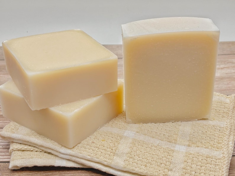 May include: Three rectangular bars of solid, off-white soap with smooth surfaces and rounded edges. The bars are stacked in a cluster, with one standing upright and two lying flat, resting on a folded, light beige, checkered fabric cloth.