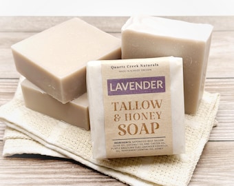 Könnte beinhalten: Drei rechteckige Stücke hellbeiger Seife. Ein Stück ist in ein beiges Papieretikett mit violettem Text eingewickelt, auf dem Lavender Tallow & Honey Soap, Quartz Creek Naturals, Made in Seaside Oregon und eine Zutatenliste stehen.