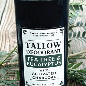 K&ouml;nnte beinhalten: Ein schwarzer, zylindrischer Deodorant-Stick mit einem wei&szlig;en Etikett, auf dem Quartz Creek Naturals, Tallow Deodorant, Tea Tree & Eucalyptus with Activated Charcoal und ein Nettogewicht von 75 g stehen. Der Beh&auml;lter hat einen Drehfu&szlig;.