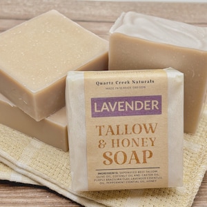 May include: Three bars of handmade lavender tallow and honey soap. The soap is wrapped in white paper with a label that reads "Quartz Creek Naturals, Made in Seaside Oregon, Lavender Tallow & Honey Soap". The label also lists the ingredients: saponified beef tallow, olive oil, coconut oil, castor oil, purple Brazilian clay, lavender essential oil, peppermint essential oil, and honey.