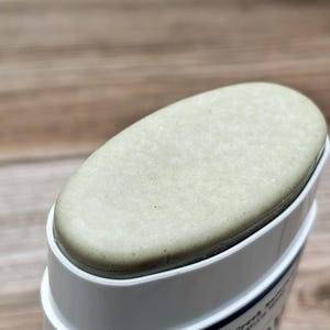 K&ouml;nnte beinhalten: Ein fester, ovaler Stift aus hellsalbeigr&uuml;nem Deodorant oder Hautpflegeprodukt in einem wei&szlig;en Kunststoffbeh&auml;lter. Die Oberseite des Produkts ist glatt und flach und ragt leicht &uuml;ber den Rand des wei&szlig;en Geh&auml;uses hinaus.