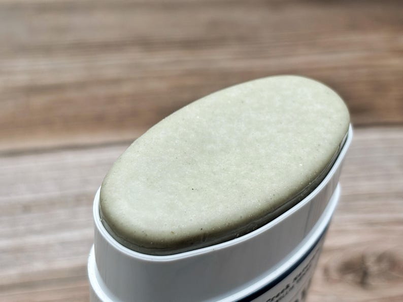 K&ouml;nnte beinhalten: Ein fester, ovaler Stift aus hellsalbeigr&uuml;nem Deodorant oder Hautpflegeprodukt in einem wei&szlig;en Kunststoffbeh&auml;lter. Die Oberseite des Produkts ist glatt und flach und ragt leicht &uuml;ber den Rand des wei&szlig;en Geh&auml;uses hinaus.
