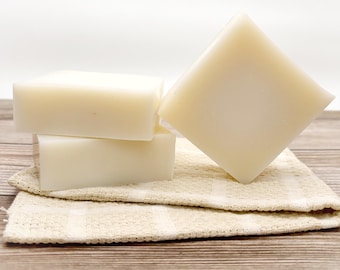 May include: Three bars of white soap stacked on top of each other on a beige and white checkered cloth. The soap is on a wooden surface.