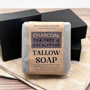 May include: Three rectangular bars of black charcoal tallow soap. The center bar is wrapped in a tan paper label with black text reading Quartz Creek Naturals, Charcoal Tea Tree & Eucalyptus Tallow Soap, and a list of ingredients.