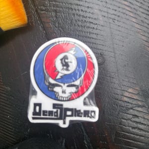 May include: A white sticker with a colorful graphic of a skull with lightning bolts and the text "Dead & Co."