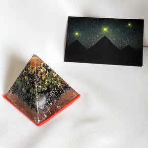 Könnte beinhalten: Eine klare Pyramide mit einer roten Basis, gefüllt mit schwarzen, grünen und goldenen Kristallen. Die Pyramide steht auf einer weißen Oberfläche, und im Hintergrund befindet sich eine Karte mit schwarzem Hintergrund und einem Sternenhimmelbild.
