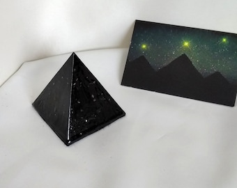 May include: A black pyramid-shaped object with a textured surface. The pyramid is sitting on a white surface. A black card with a starry night sky and three green stars is in the background.