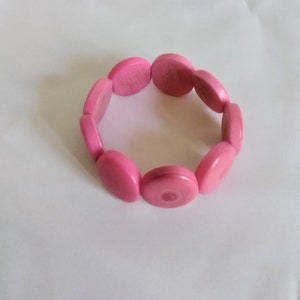 May include: A pink bracelet composed of oval-shaped beads. The beads are a solid, vibrant pink color and are strung together to form a circular shape. The bracelet is shown against a white background.