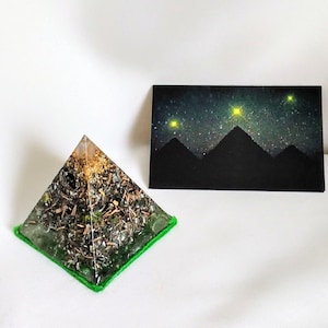 Pode incluir: Uma pirâmide transparente com uma base verde, cheia de vários cristais e flocos dourados. A pirâmide está sobre uma superfície branca, com um cartão preto atrás. O cartão tem um fundo azul escuro com um céu estrelado e três montanhas, com três estrelas amarelas acima das montanhas.