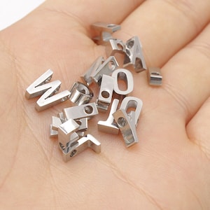 May include: A collection of silver metal letter and number charms. The charms are small and have a hole for stringing.