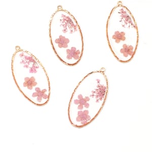 May include: Four oval pendants with gold-coloured frames. Each pendant features dried pink flowers set against a clear background. The pendants have a small loop at the top for hanging.
