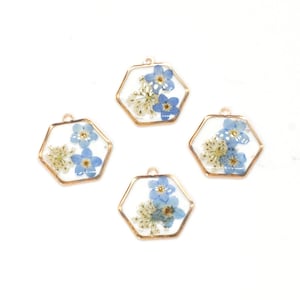 May include: Four hexagonal pendants with gold-colored frames. Each pendant contains a cluster of small blue and white dried flowers set in clear resin. The pendants are designed for jewelry making or crafting.