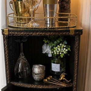 May include: A black and gold bar cabinet with a glass top, holding barware and a bottle of champagne in a gold bucket. The cabinet has a lower shelf with a wine glass, flowers, and a decorative silver container. A lamp sits on top.