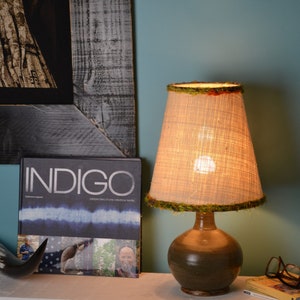 Op de afbeelding: Een bruine keramische tafellamp met een lampenkap van geweven stof. De lampenkap heeft een groene en bruine rand. De lamp staat op een tafel naast een boek met de titel "Indigo" en een bril.