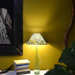 Op de afbeelding: Een tafellamp met een groene glazen flessenvoet en een lampenkap met een tropisch bladmotief. De lamp staat op een tafel met een stapel boeken en een groene plant.