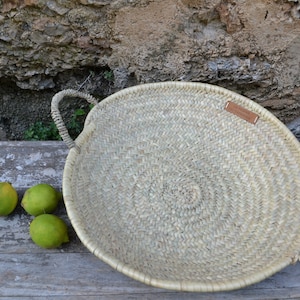 Pode incluir: Uma cesta grande, redonda e tecida com duas alças. A cesta é feita de materiais naturais e tem uma cor castanho claro. A cesta está sentada sobre uma superfície de madeira com três limões verdes à sua frente.