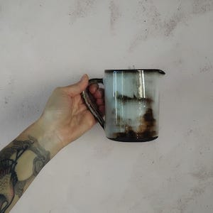Op de afbeelding: Een keramische kan met een handvat, met een lichtblauwe en bruine glazuur. De kan heeft een donkerbruine rand en basis. Het ontwerp heeft een rustieke, handgemaakte uitstraling.