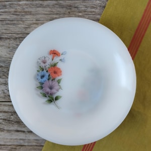 Könnte beinhalten: Weißer, runder Teller mit floralem Design. Die Blumen sind in Orange-, Blau-, Lila- und Weißtönen gehalten, mit grünen Blättern. Der Teller hat einen leicht erhöhten Rand.