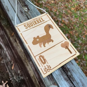May include: Wooden sign with the word "SQUIRREL" at the top, featuring a pixelated squirrel illustration. The sign has a light brown border and a white string for hanging. A heart and paw prints are also included.