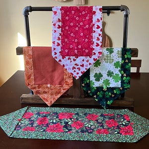 May include: Three table runners displayed on a wooden stand. The runners are made of fabric with different patterns: red and white hearts, green shamrocks, and red poinsettias.