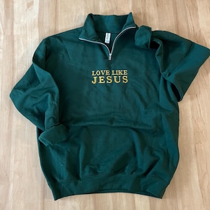 Könnte beinhalten: Ein dunkelgrüner Pullover mit Reißverschluss und einer goldenen Stickerei mit der Aufschrift "LOVE LIKE JESUS".