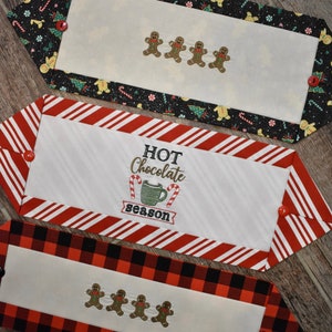 May include: Three white fabric table runners with different holiday-themed embroidery designs. The top runner has a black and white background with gingerbread men. The middle runner has a red and white striped background with the text "HOT Chocolate season". The bottom runner has a red and black plaid background with gingerbread men.