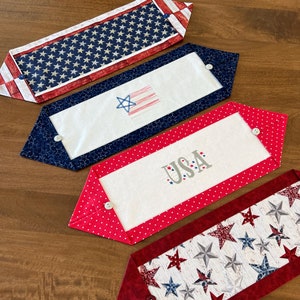 May include: Four patriotic table runners with red, white, and blue designs. One features a field of stars, another has a star and stripe design, and a third displays the word "USA". Each runner has a button closure and is set on a wooden surface.