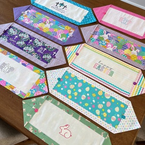 May include: A set of eight Easter table runners with various designs. The runners feature bunnies, eggs, crosses, and the words "Happy Easter", "He is Risen", and "Easter Blessings".