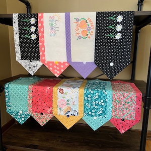 May include: A set of six colorful table runners with different patterns, including floral, polka dot, and geometric designs. The runners are displayed on a wooden shelf with black metal supports. The runners are all trimmed with a white fabric and have a button detail.