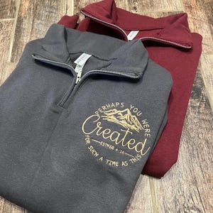 Könnte beinhalten: Zwei Sweatshirts mit Viertelreißverschluss, eines in Anthrazitgrau und eines in Burgunderrot, werden auf einer Holzoberfläche präsentiert. Das graue Sweatshirt hat eine goldfarbene Stickerei mit dem Text "Perhaps You Were Created For Such A Time As This."