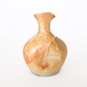 May include: A small, round, ceramic vase with a brown and orange glaze. The glaze has a drip effect, creating a unique and rustic look.