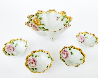 May include: A set of four porcelain bowls with a floral design. The bowls are white with a gold rim and feature pink and yellow roses with green leaves. The largest bowl has three legs.