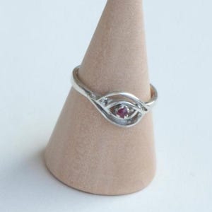Könnte beinhalten: Ein silberner Ring mit einem filigranen Design, das einen kleinen rosafarbenen Edelstein zeigt. Der Ring wird auf einem hölzernen Ringständer präsentiert.