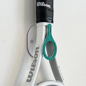 May include: A white and gray Wilson tennis racket is mounted on a wall with a teal-colored holder. The racket has a black grip and the Wilson logo is visible. The wall mount is white with a purple accent.