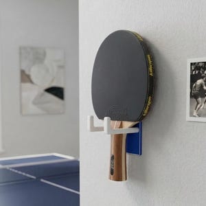 Może przedstawiać: Czarna i żółta rakietka do tenisa stołowego Killerspin zamontowana na białym i niebieskim uchwycie ściennym. Rakieta ma drewniany uchwyt i jest eksponowana na białej ścianie z oprawionym czarno-białym zdjęciem.