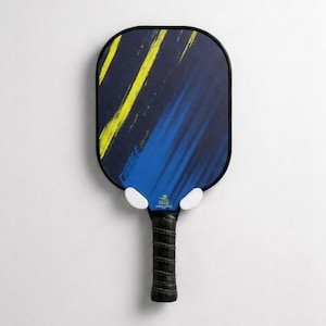 May include: A pickleball paddle with a black frame and a blue and yellow design. The paddle has a black grip and is mounted on a white wall. The word "CHARGE" is visible on the paddle.
