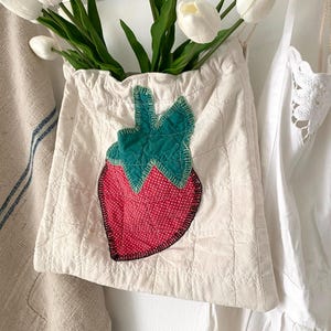 May include: A cream-colored drawstring bag with a quilted pattern and a large strawberry applique. The strawberry features a red dotted center, green leaves, and a black outline. The bag is filled with white tulips.