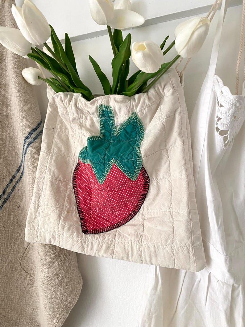 May include: A cream-colored drawstring bag with a quilted pattern and a large strawberry applique. The strawberry features a red dotted center, green leaves, and a black outline. The bag is filled with white tulips.