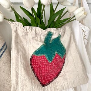 May include: A cream-colored, quilted fabric bag with a large, appliqu&eacute;d strawberry design. The strawberry is red with white polka dots and green leaves. The bag is filled with white tulips.