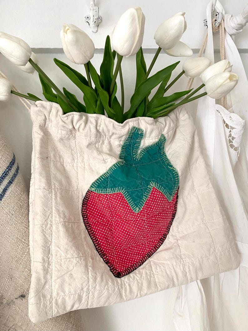 May include: A cream-colored, quilted fabric bag with a large, appliqu&eacute;d strawberry design. The strawberry is red with white polka dots and green leaves. The bag is filled with white tulips.